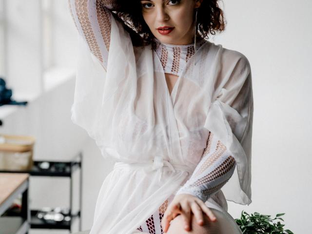 CurlyVixen with curly red hair, wearing a sheer white dress, showing her tits through the fabric, arm raised, hand on knee, looking at camera, in a bright room.