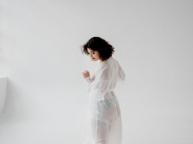 CurlyVixen with curly dark hair, wearing a sheer white robe, looking down, revealing blue floral-patterned panties and a subtle view of her ass. White background.