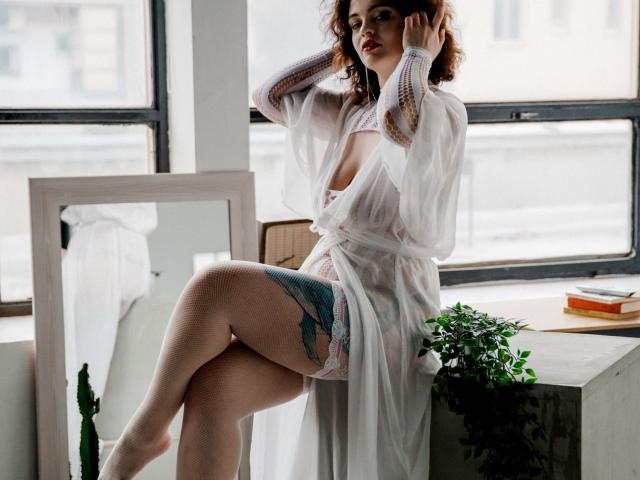 CurlyVixen with curly brown hair, wearing a sheer white robe, exposing her tits and a colorful thigh tattoo, sits by a window, legs crossed, looking seductive.