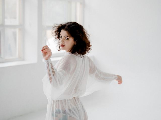 CurlyVixen with curly brown hair, wearing a sheer white dress, stands in a bright room, her back to the camera, slightly turning to look over her shoulder.