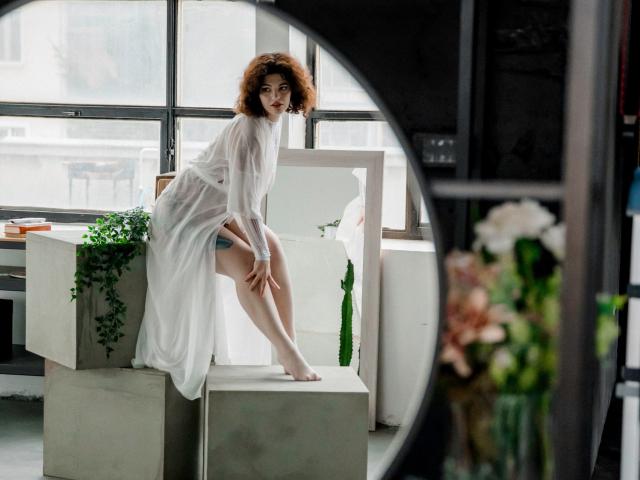 CurlyVixen with curly red hair, white sheer dress, sitting on a concrete block, legs bent, showing her bare ass, in a bright, modern room with plants.