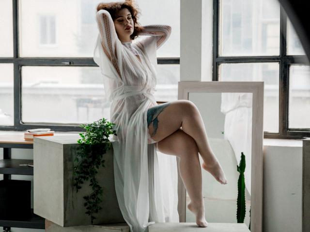 CurlyVixen with curly brown hair, wearing a sheer white dress, sits on a table, legs crossed, showing a tattoo on her thigh, hands in her hair, looking sultry.