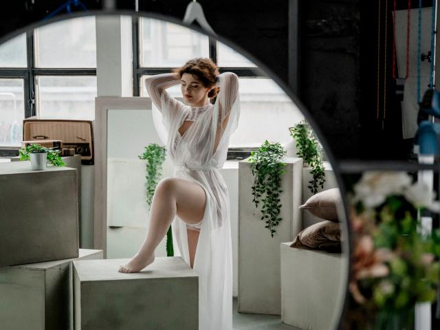 CurlyVixen with short brown hair, wearing a sheer white dress, stands on a gray block, one leg up, hands behind head, showing her white panties and bare legs.