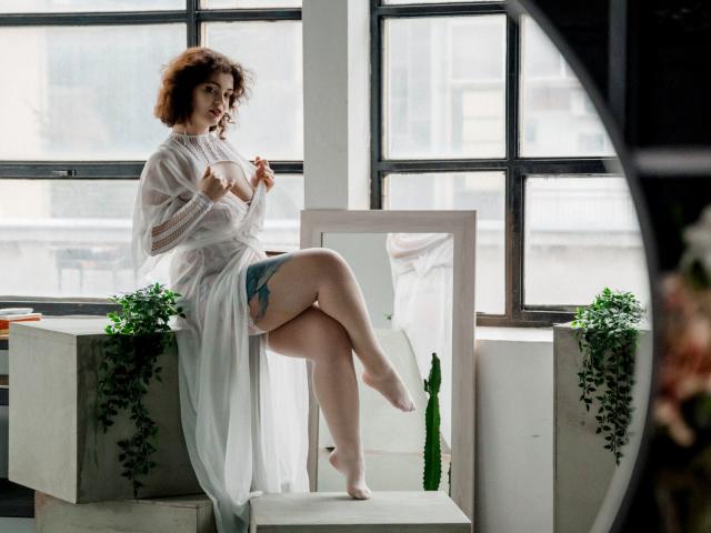 CurlyVixen with curly brown hair, wearing a sheer white robe, exposing her tits and a blue tattoo on her thigh, sits on a white chair, legs crossed.