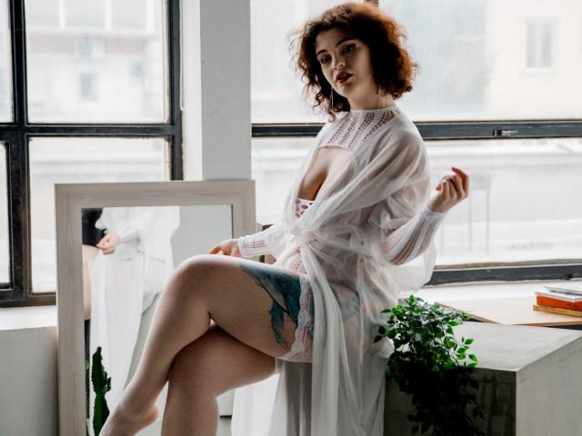 CurlyVixen with curly red hair, pale skin, and a tattoo on her thigh, wearing a sheer white dress, revealing her tits and ass, sitting by a window, looking at camera.