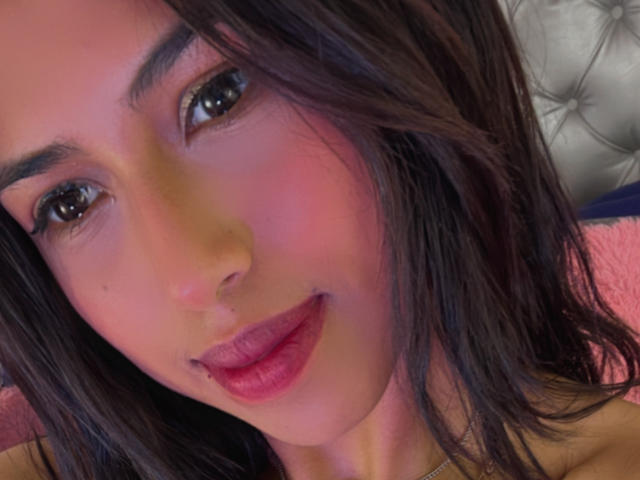 MyaaTyler's face with dark brown hair, pink blush, and red lips, looking at the camera with slightly parted pink lips. She has a small nose and dark eyes.