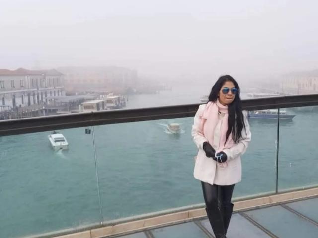 SaraLunaBella69 with long black hair, wearing a pink scarf, white coat, black gloves, and blue sunglasses, standing on a balcony with glass railing, boats in teal water.