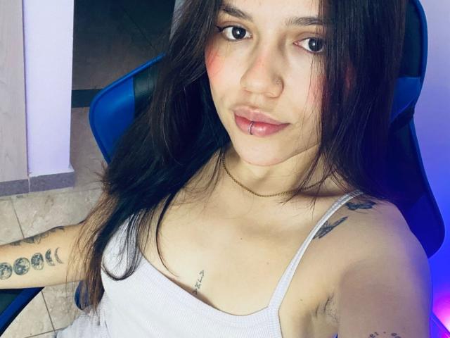SophiaMarx with long brown hair, pink lipstick, white tank top, tattoos on arm and chest, nose ring, sitting in blue chair, slight smile, visible cleavage, light skin, blue light.