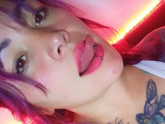 SweetAmeliee met paars haar, bruine ogen en een roze tongpiercing, die haar tong uitsteekt. Ze heeft een vlindertatoeage op haar borst. Haar lippen zijn glanzend en vol..