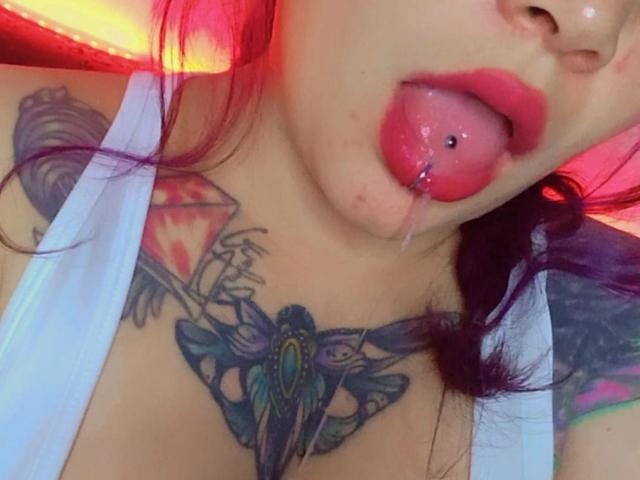 SweetAmeliee with tongue out, tongue pierced, drooling, red lipstick, purple hair, white tank top, tattoos on chest and shoulder, visible cleavage, colorful tattoos, slightly angled headshot.