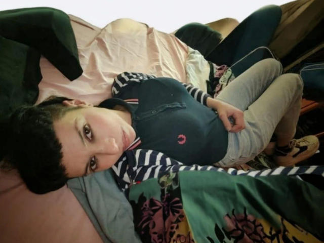 LadyRiot-hot with short black hair, lying on a bed, wearing a black and navy polo with striped sleeves, gray pants, and beige shoes, looking up at the camera.