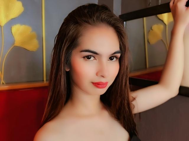 Kirsten, mit nassen, braunen Haaren, roten Lippen und heller Haut, steht oben ohne, ein Arm erhoben, und blickt in die Kamera. Sie befindet sich in einem warm beleuchteten Raum, im Hintergrund sind Gemälde mit gelben Blumen zu sehen..