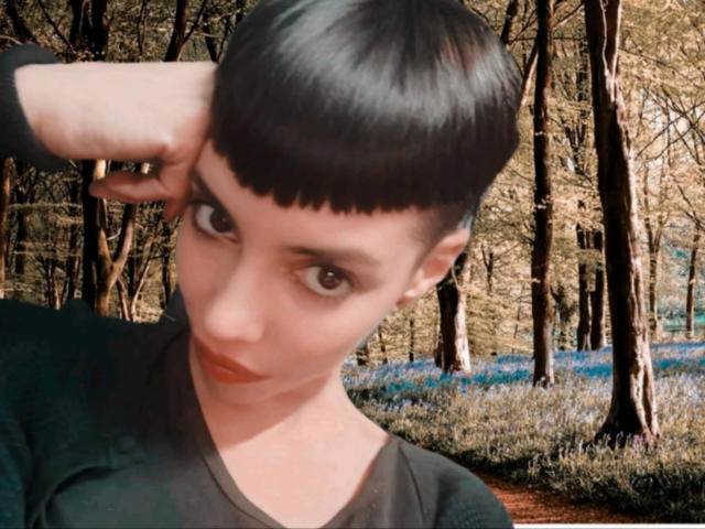 LadyRiot-hot with a black bob, raising her right arm, wearing a black shirt, looking at the camera with large brown eyes, standing in a forest with blue flowers.