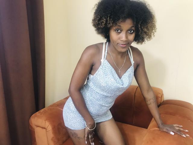 LuceLaJolie with dark skin and curly hair, wearing a blue patterned dress, kneeling on an orange couch, showing tattoos on her thighs, with a bracelet and necklace.