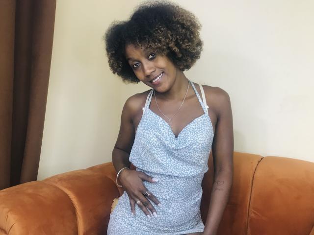 LuceLaJolie, a cute dark-skinned woman with curly brown hair, wearing a blue patterned dress, smiling, hand on her belly, sitting on an orange couch.