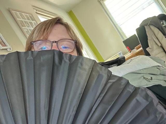Chantallsweat-hot with glasses, blue eyes, and brown hair, holding a black fan over their face, in a bright room.
