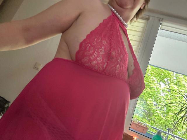 Chantallsweat-hot in a red lace halter babydoll, showing side boob, with a pearl necklace, and a pink sheer lower part, standing near a window with green trees outside.