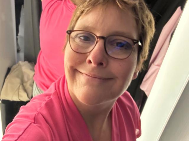 Chantallsweat-hot, short blonde hair, glasses, pink shirt, smiling slightly, light skin, subtle wrinkles, medium-sized nose, visible cheek creases, slight double chin, casual selfie, upper body shot.