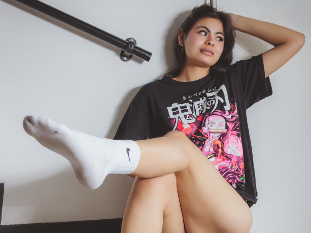 CrystallThonpsom with dark hair, wearing a black t-shirt, white socks, and no pants, sitting with one leg raised, showing her bare ass and a confident smile.