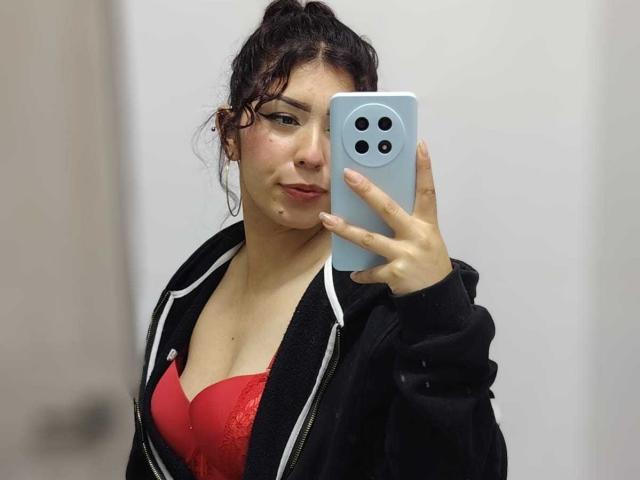ToryPussCaty, con cabello oscuro y rizado, vestida con un sostén de encaje rojo y una chaqueta negra con cremallera, se toma un selfie frente al espejo con un teléfono azul, mostrando sus pechos..