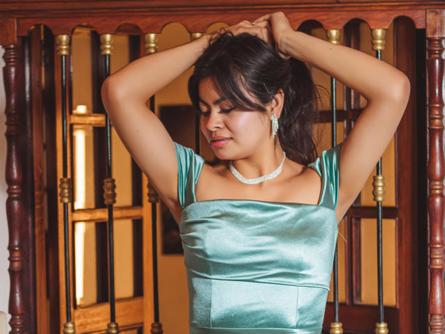 CrystallThonpsom with arms raised, wearing a shiny green satin dress, silver earrings, and a matching necklace, standing in front of a wooden railing. Her eyes are closed.