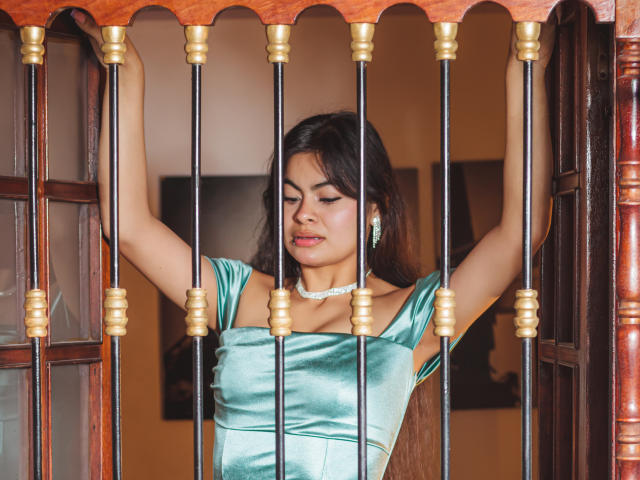 CrystallThonpsom with arms up, green satin dress, gold earrings, white necklace, standing behind black and gold barred window, brown wooden frame, looking down, brown hair, soft smile.