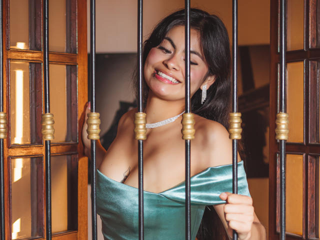 CrystallThonpsom with medium tits, smiling, green off-shoulder dress, diamond necklace, and earrings, holding metal bars, dark hair, brown skin, and pink lipstick.