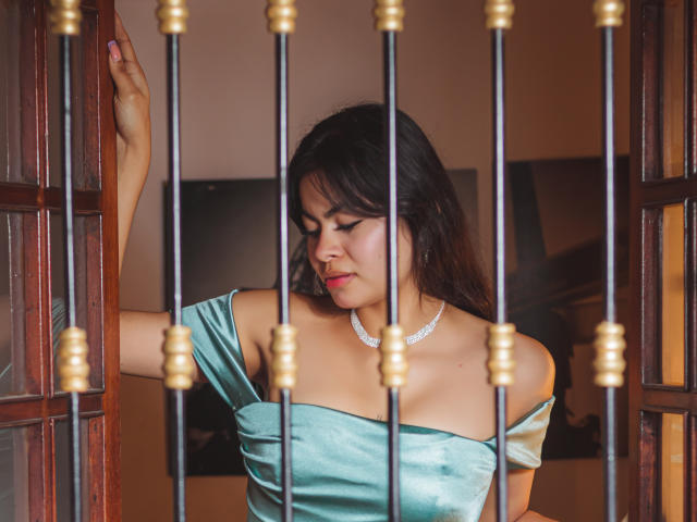 CrystallThonpsom with dark hair, medium tits, wearing a shiny green off-shoulder dress, silver necklace, standing by a wooden door, touching the frame, looking down, photo.