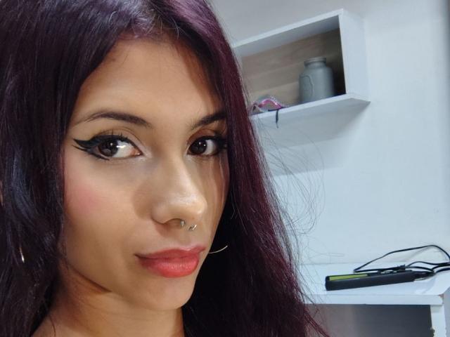 DanikaSerov with dark purple hair, black eyeliner, nose piercing, red lipstick, and hoop earrings, smiling slightly, holding a phone in a white-shelf room.