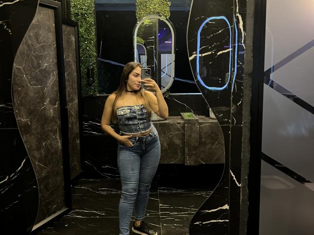 SharonKlart with long brown hair, strapless denim top, and tight blue jeans, taking a mirror selfie in a black marble room, holding her phone, smiling.