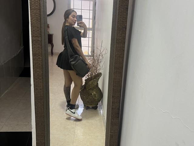 SharonKlart, con un minuscolo vestito nero, scarpe da ginnastica bianche e calze nere, che tiene una borsa nera, mostra il sedere e i tatuaggi, mentre scatta un selfie allo specchio..