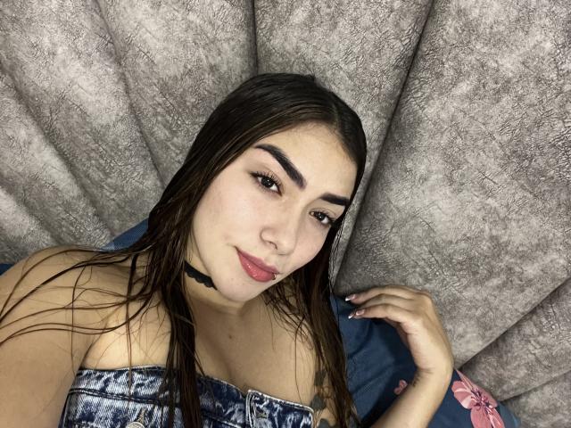 SharonKlart with wet brown hair, dark eyebrows, pink lipstick, and a blue strapless top, lying on a gray textured surface, wearing a black choker, and showing her tits.