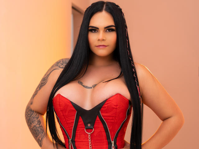 JassmineTS with long black hair, red and black corset, large boobs, tattooed arm, chain detail, pink lipstick, confident expression, standing, hands on hips.