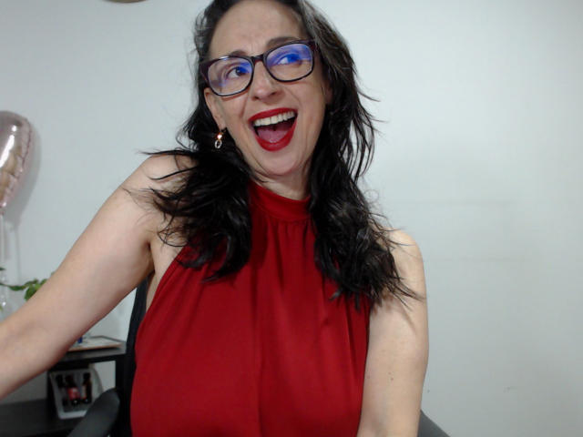 AryanaBurning with wild black hair, blue glasses, red sleeveless top, and red lipstick, smiling open-mouthed, showing her tits. She's sitting with a light gray background.