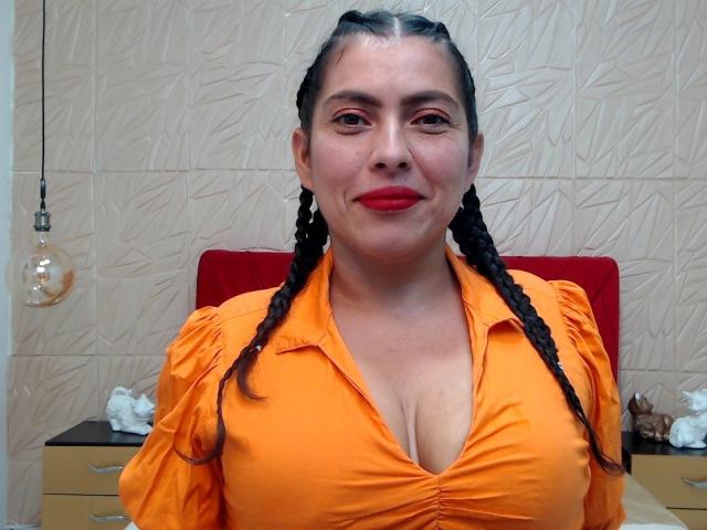 Luciana Montiel, con i capelli neri intrecciati, una camicia arancione con bottoni, rossetto rosso e scollatura visibile, sorride leggermente, in piedi di fronte a un muro con una lampadina appesa..
