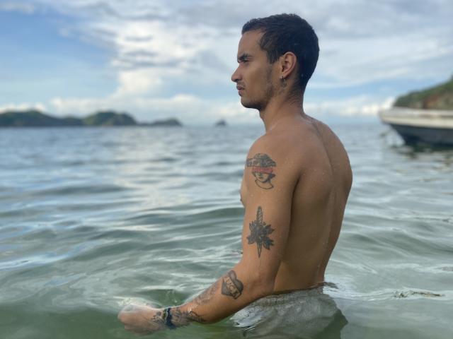 LailaAsher, shirtless, standing in green water, showing tattoos on arms and back, short dark hair, looking right, small boat in background, cloudy sky, medium skin, muscular build.