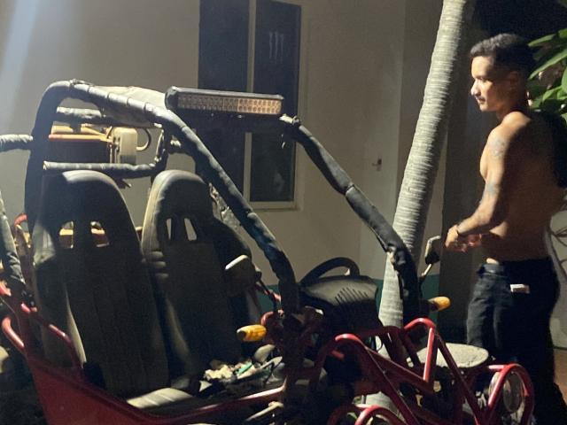 Shirtless LailaAsher stands next to a red, damaged, off-road vehicle with torn black fabric and two gray seats, holding a tool, with a palm tree to the right.