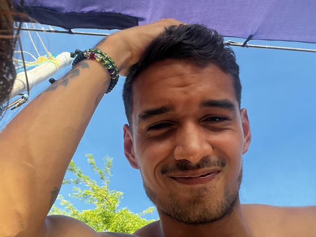 LailaAsher, shirtless, with short dark hair, tattoos on arm, green bead bracelet, smiling under purple umbrella, clear blue sky, hand in hair, slight beard, tan skin, cute, attractive.