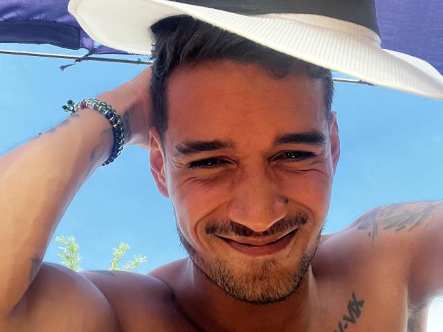 LailaAsher è un uomo abbronzato, senza camicia, che sorride e indossa un cappello da sole bianco. Ha la barba, dei tatuaggi e un braccialetto, con un cielo blu sullo sfondo..