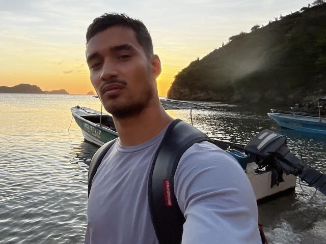 LailaAsher with short dark hair, beard, wearing a gray shirt and black backpack, standing by small boats at sunset, yellow-orange sky, calm water, and rocky hillside in background.