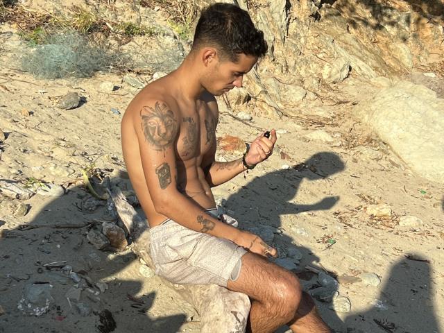 LailaAsher, shirtless, with tattoos on chest and arm, sitting on sandy beach, wearing beige shorts, holding black object, looking down, sunlight casting shadow.