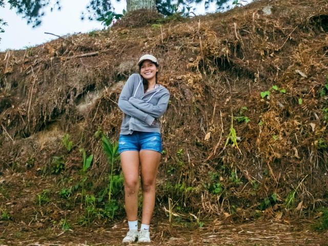 Laila Asher steht mit verschränkten Armen da, trägt eine graue Kapuzenjacke, eine weiße Mütze, blaue Shorts und weiße Turnschuhe, lächelt und hat einen braunen Hügel aus Baumwurzeln im Hintergrund..