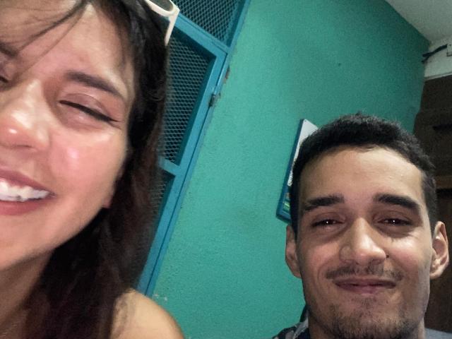 LailaAsher tiene el cabello castaño, los ojos cerrados y una sonrisa que muestra dientes blancos. El amigo masculino de LailaAsher tiene el cabello corto y negro, un bigote y una sonrisa burlona. Ambos están en una habitación con paredes color turquesa..
