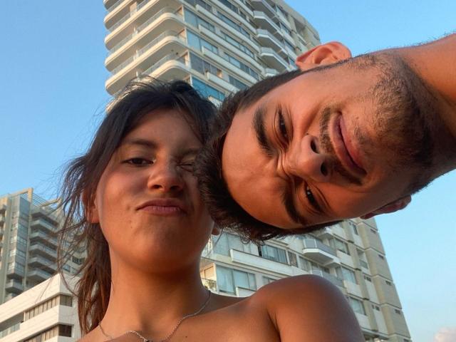 LailaAsher with a cute, slightly smug expression, and a guy with a mustache and short hair, both shirtless, in front of a tall apartment building, under a clear blue sky.