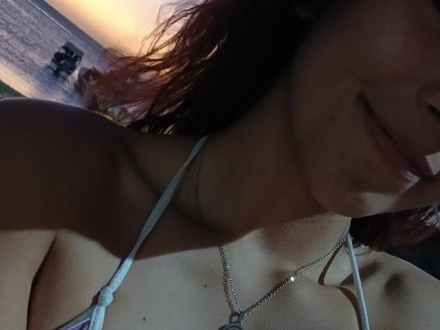 LailaAsher's upper body, showing her bare shoulders and white halter top straps. She's wearing a necklace, with her face partially in the frame, smiling. Her tits are visible.