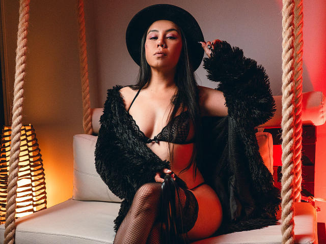 CharlotteJoliie with long black hair, wearing a black hat, lace bra, and fishnet stockings, sitting on a swing, covered in a fluffy black jacket, showing her tits and ass.