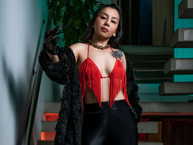 CharlotteJoliie with red fringed top, black furry coat, black leggings, and black lace gloves, standing by stairs, showing a tattoo on her right shoulder, looking confident.