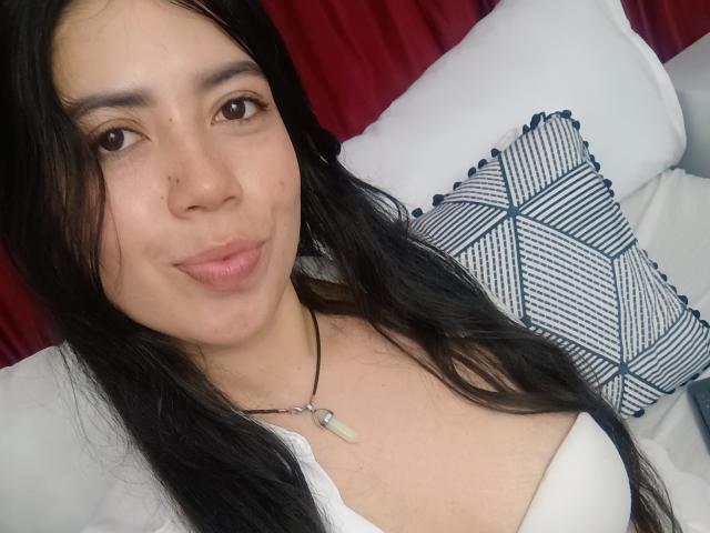 CristalDuport with long black hair, brown eyes, light skin, small nose, pouty lips, white top, necklace with green stone, and blue-striped pillow in the background.