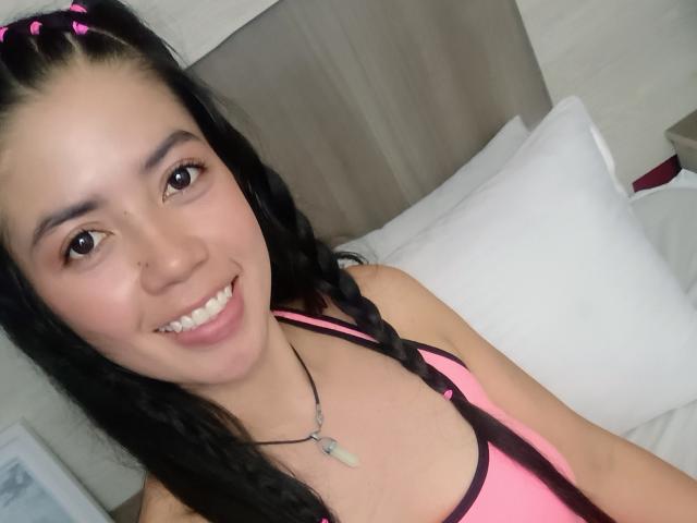 CristalDuport with black braids, pink top, and necklace, smiling at the camera, showing her tits, brown eyes, and cute face. She has pink hair ties in her braids.