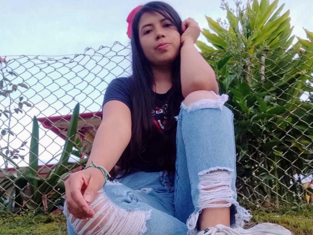 CristalDuport with long black hair, wearing a red flower in her hair, black t-shirt, and ripped blue jeans, sitting outdoors with one knee up, pouty expression, green bracelet.
